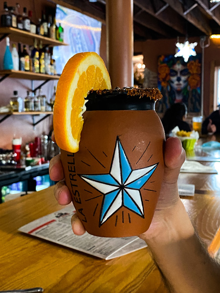 Asheville tacos Ay Caramba Mexican Grill with white handing holding up drink in clay glass with white and blue star and garnished with an orange