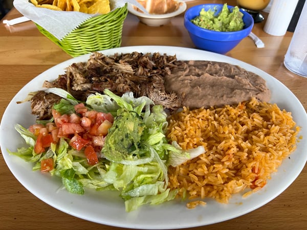 Ay Caramba Restaurant in Asheville Carnitas on white plate with lettuce, tomato, orange colored rice, and refried beans with chips and guacamole in the background
