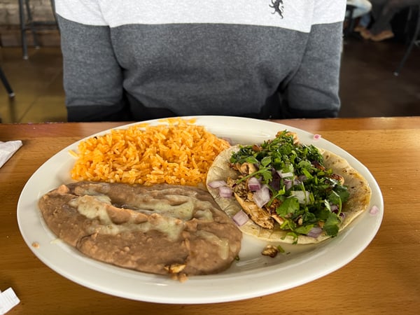 Ay Caramba Restaurant Tacos in Asheville, NC with white plate filled with brown refried beans, soft taco filled with meat and cilantro, and orange rice in front of person wearing an ombre gray sweater