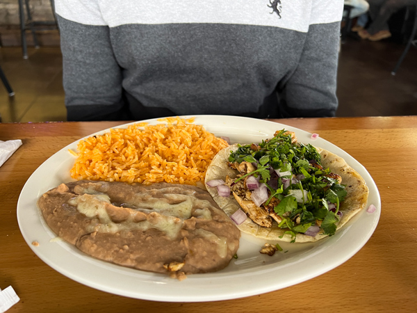 Ay Caramba Restaurant Tacos in Asheville, NC with white plate filled with brown refried beans, soft taco filled with meat and cilantro, and orange rice in front of person wearing an ombre gray sweater