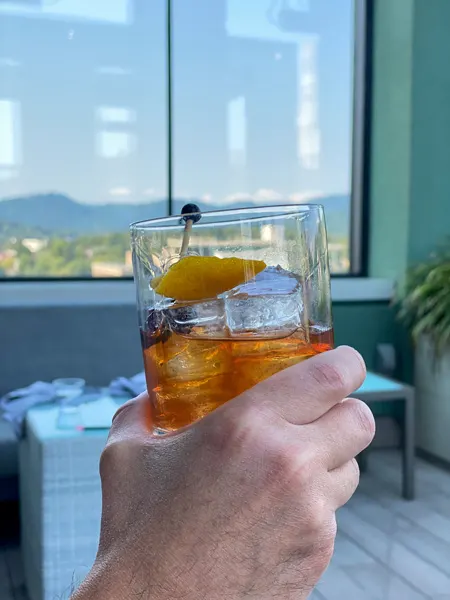 Best Rooftop Bars Asheville Capella On 9 with white hand holding up an Old Fashioned Craft cocktail in front of window with view of Blue Ridge Mountains