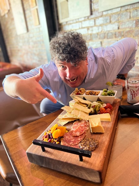 Bottle Riot Wine Bar Asheville North Carolina with white male with white gray curly hair pointing at a huge charcuterie board filled with olives, meats, and cheeses
