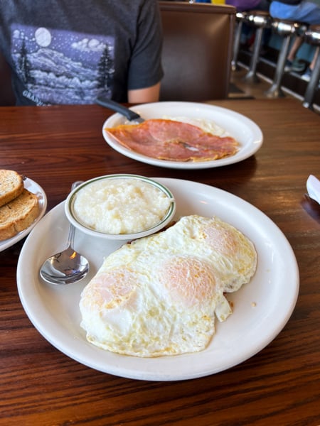 Five Points Restaurant Asheville Breakfast with two plates on brown table, one with three over easy eggs and grits and one with pink ham and over easy egg with toast on side