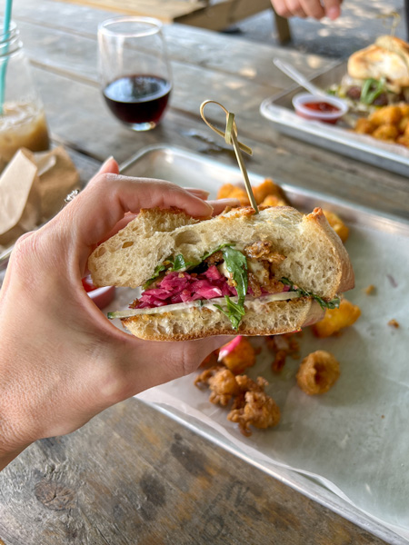 Haywood Common Fried Clam Sandwich Lunch Asheville NC with white hand holding up sandwich with tater tot side and wine in background on picnic table