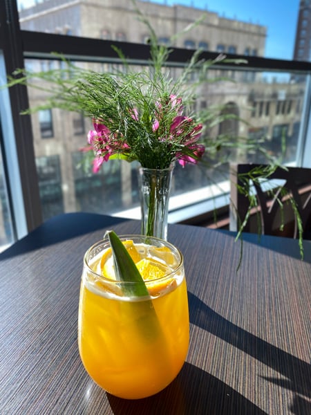 Hemingways Cuba Asheville NC yellow mango cocktail on brown table top garnished with green veggie stick and small vase of flowers