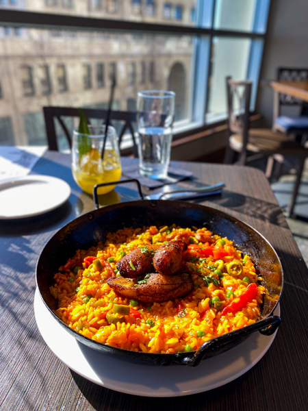 Hemingways Cuba Rooftop Restaurant Asheville with bowl of yellow rice, chicken, and fried plantains with peppers and green olives on table with cocktails and water overlooking Grove Arcade