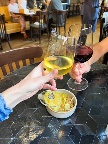 La Bodega by Curate Wine Bar Asheville NC with white male hand clinking red wine glass with white woman's white wine glass and olive tapas between them on table