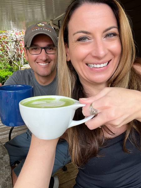 Liberty House Cafe in Asheville, NC with white brunette woman with blonde highlights holding green tea matcha latte doing a cheers with a white brunette male in hat and glasses holding blue mug of coffee