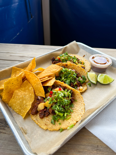 Mamacitas Taco Temple in Asheville NC Tacos on brown chef's paper on tin plate with tortilla chips on side and topped with lettuce, corn, and cilantro. There is a lime garnish on the side along with takeaway dipping sauce