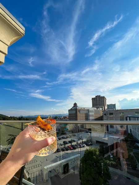 White hand holding cocktail in clear low ball glass up over rooftop with mountains, buildings, and blue sky with clouds in the background at Pillar Rooftop Bar in Asheville, NC