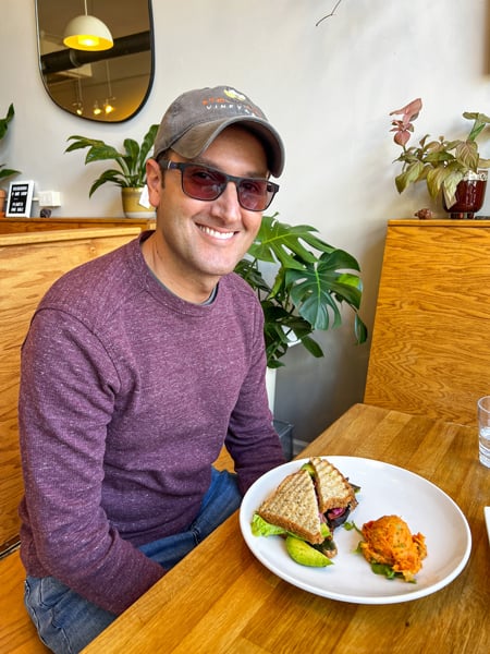 Tom, a white brunette male in maroon sweater with hat and sunglasses, with gluten free sandwich on plate with sweet potato salad at Pulp and Sprout in Asheville NC 