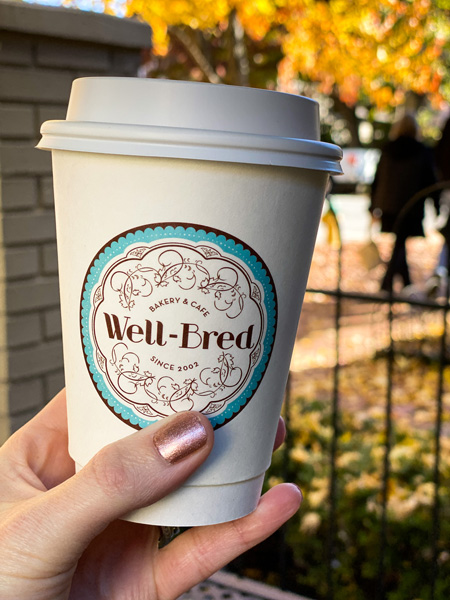 Well Bred Bakery and Cafe coffee in takeaway cup held up by white hand with pink nails in front of fence and fall foliage trees