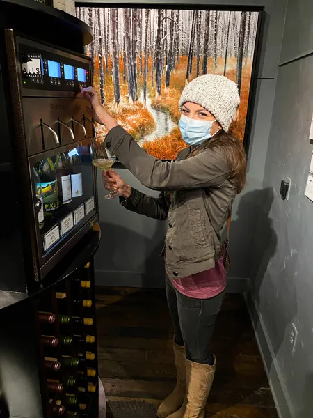 Wine Dispense Walls Wine Bar Asheville NC with white woman in green jacket, white hat, jeans getting wine