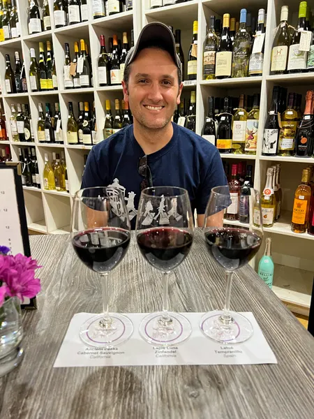 Wine Sage and Gourmet Hendersonville NC with white male in hat and t-shirt with three glasses of red wine in front of him on gray table surrounded by wine bottles on shelves
