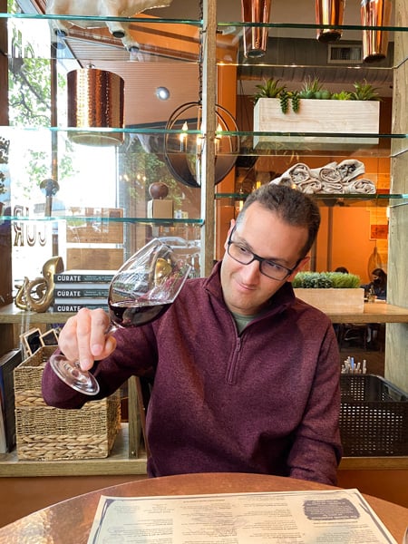 Tom, a white brunette male in a red sweater, swirling a glass of red wine and looking at it at Curate Restaurant in Asheville, NC