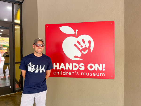 Hands On Children's Museum Hendersonville NC with image of white brunette male with sunglasses and hat in front of their red sign with apple and hand print