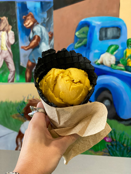 Christine's white hand holing up a vegan black waffle cone filled with vegan pumpkin ice cream in front of a mural at The Hop Ice Cream in Asheville, NC