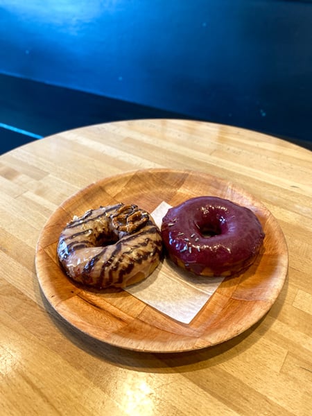 Vortex Doughnuts Asheville NC with two donuts on brown plate with brown table and blue wall background.  One donut in blueberry glazed and the other is chocolate with a pretzel on top