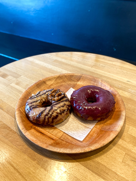 Vortex Doughnuts Asheville NC with two donuts on brown plate with brown table and blue wall background. One donut in blueberry glazed and the other is chocolate with a pretzel on top