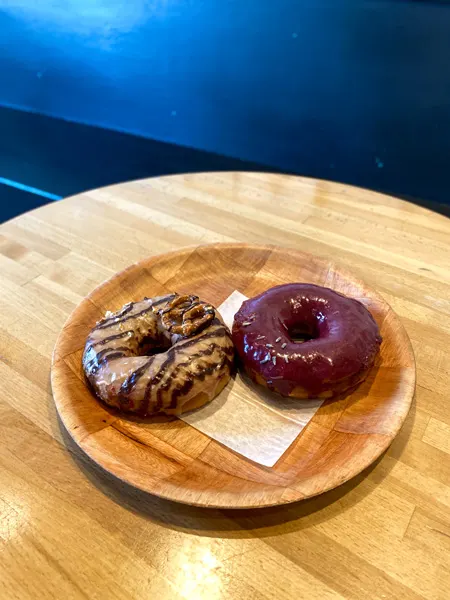 Vortex Doughnuts Asheville NC with two donuts on brown plate with brown table and blue wall background. One donut in blueberry glazed and the other is chocolate with a pretzel on top