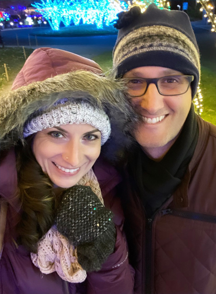 Winter Lights in Asheville NC Selfie with white brunette male and female in winter coats with warm knit winter hats and light displays in the background