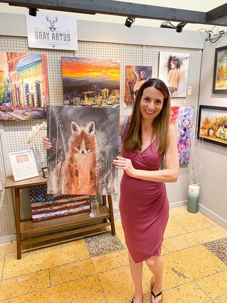 Woolworth Walk in Asheville artist stall with paintings on the wall and white brunette female in pink dress holding a painting of a fox for purchase