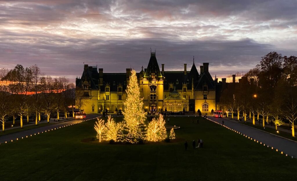 Biltmore House at Christmas lit up at night with holiday trees out front and sunset over the Blue Ridge Mountains in Asheville, NC