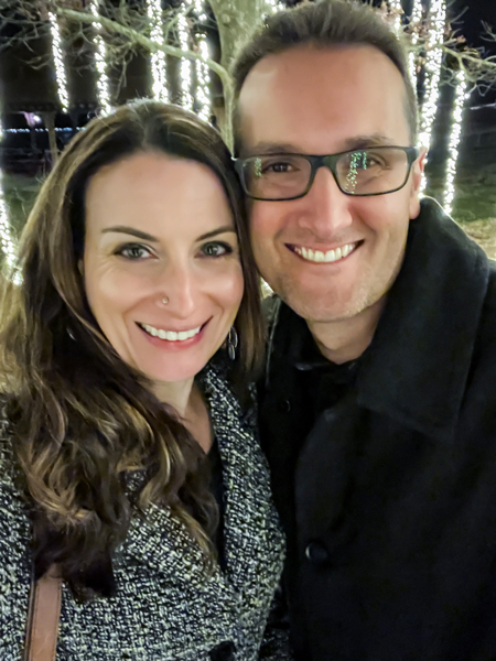 Christine and Tom, a white brunette male and female, in winter peacoats in front of yellow lights hanging from trees at Biltmore Estate at Christmastime