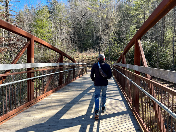 DuPont State Forest Hooker Falls Access Area with man in fleece and hat walking away from camera across a brown bridge over water