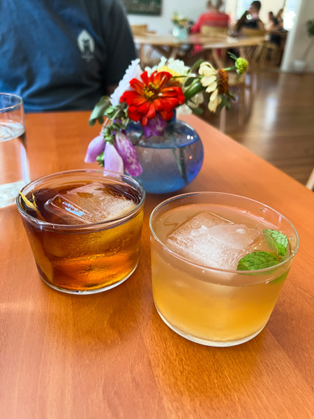 ELDR Restaurant Asheville Brunch Cocktails with brown cocktail and yellow cocktail with green garnish and giant square ice cube on brown table with vase of flowers in the background