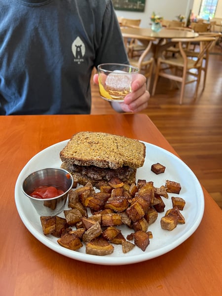 ELDR Restaurant Asheville Burger on gluten free bread with fried fingerling potatoes on white plate on brown table with person holding up cocktail
