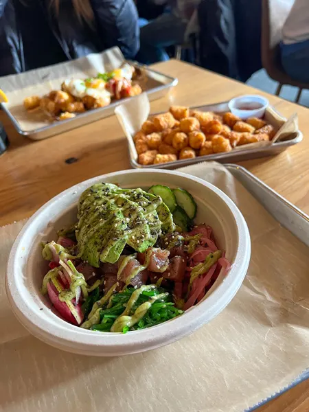 Haywood Common Asheville Brunch poke bowl in take away container with tuna, avocado, white ice, seaweed, and green herbs with tator tots and egg breakfast platter blurred in background