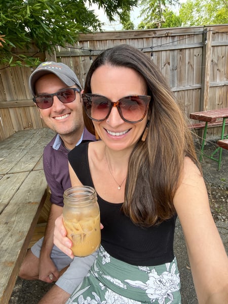 Christine and Tom, a white brunette male and female both wearing sunglasses outside on picnic table; Christine is holding an iced latte in a mason jar; they are at Haywood Common in Asheville, NC on the outdoor patio