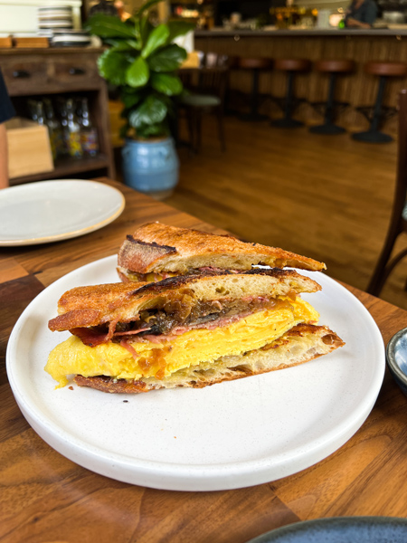 La Bodega by Curate Brunch Asheville North Carolina sandwich with egg, meats, and sautéed onions on white plate on brown table