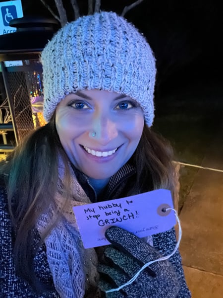Christine, a white brunette female in a winter hat and coat holding a hand-written ornament that says, "my hubby to stop being a grinch" to hang on the wishing tree at Winter Lights at the NC Arboretum in Asheville