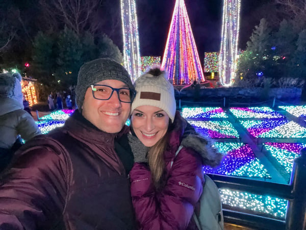 50-Foot Tree of Lights behind the lighted Quilt Garden with Christine and Tom, a white brunette male and female in winter hats and coats, taking a selfie at Winter Lights at The NC Arboretum in Asheville, North Carolina