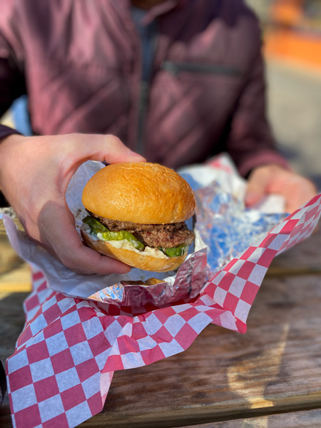 Baby Bull Burgers Asheville NC with man in purple coat holding up a burger in gluten-free bun over basket on outdoor picnic table