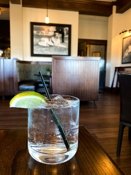 Biltmore Estate Cedrics Tavern Cocktail Asheville NC with gin and tonic garnished with lime on brown table and black and white photo of Cedric on wall in background