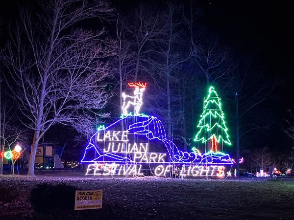 Lake Julian Park Festival of Lights entrance sign with Christmas light display including a tree and reindeer on a blue mountain