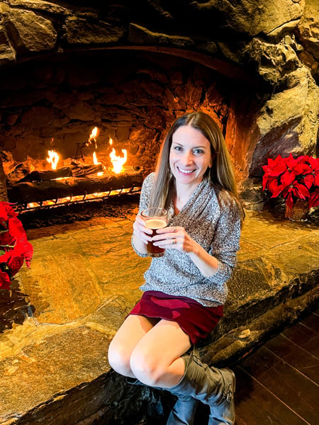 Christine, a white brunette female in red skirt, gray boots, and gray sweater sitting with beer in front of fireplace at Edison restaurant at The Omni Grove Park Inn