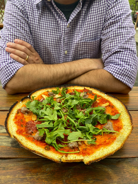 All Souls Pizza River Arts District Asheville with man in front of gluten-free polenta crust cheeseless pizza with red sauce, arugula, and roasted garlic; his arms are crossed and he's wearing a purple and white stripped shirt