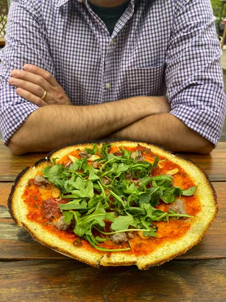 All Souls Pizza River Arts District Asheville with man in front of gluten-free polenta crust cheeseless pizza with red sauce, arugula, and roasted garlic; his arms are crossed and he's wearing a purple and white stripped shirt