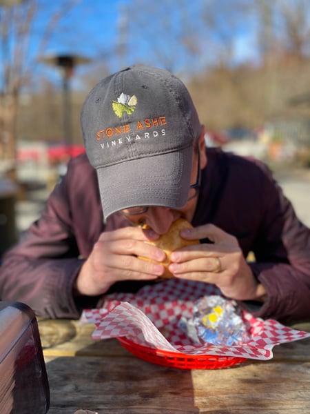 Tom, a white brunette male in purple coat and gray hat, eating burger from red basket at Baby Bull in the River Arts District in Asheville, NC