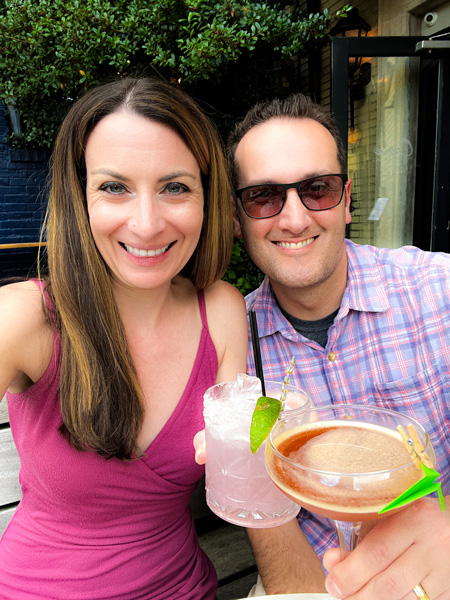 Christine and Tom, a white brunette female in a pink dress and a white brunette male in plaid shirt and sunglasses, taking a selfie doing a cheers with cocktails at Crave Dessert Bar in Asheville, NC