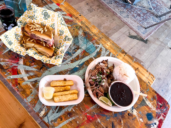 Guajiro Cuban Comfort Food Asheville NC Food Truck with takeaway plates filled with pork and bean platter, a side of yucca fries, and a Cuban sandwich on a wooden picnic table inside Great Wild Nowhere Art Bar