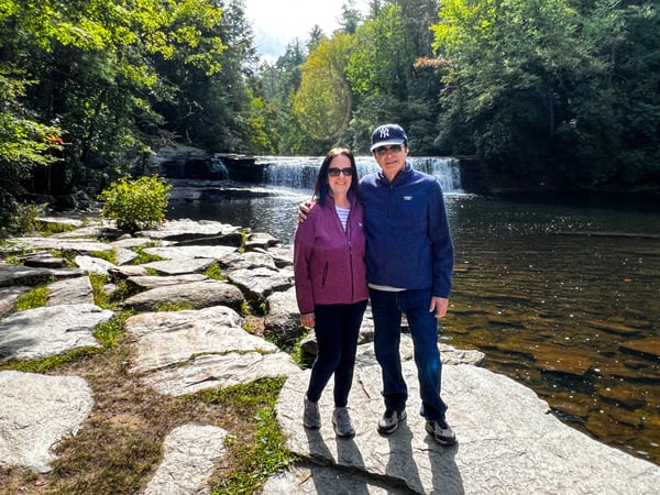 Christine's mom and dad, a white brunette male and female in blue and maroon hoodies, in front of Hooker Falls at DuPont State Forest (one-tier waterfall)