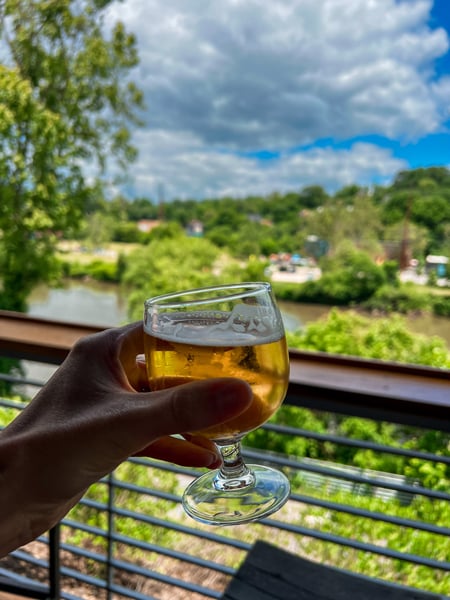 New Belgium Brewery in Asheville NC with white hand holding up a yellow lager in beer glass with French Broad River blurred in the background and blue cloudy sky