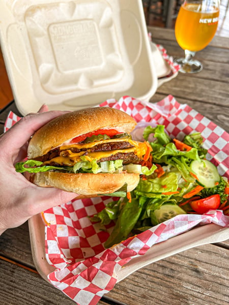 White hand holding up a Vegan Burger paired with a salad in a takeaway container from the Smokin Onion Food truck at New Belgium Brewing Company in Asheville, NC