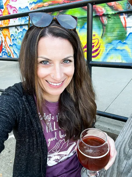 Wedge Brewery and Chop Shop Food Truck River Arts District Restaurants, Asheville with white brunette woman in dark gray zip up hoodie and purple shirt sitting at a picnic table with a craft beer with sunglasses on her forehead and mural on building in background