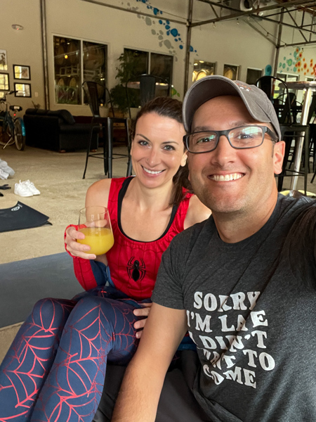 Christine and Tom, a white brunette female in Spiderman leggings and white brunette male with mimosa, on yoga mats for Halloween yoga at pleb urban winery in Asheville, NC